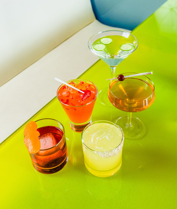 A vibrant green table displays colorful cocktails: an orange drink, a clear one with a garnish, a yellow drink with a sugar rim, a blue martini, and an orange drink with a cherry.