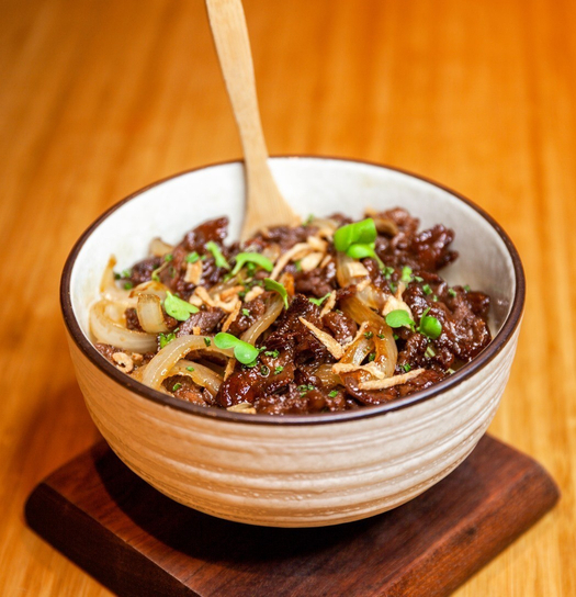 Bowl of noodles with ground meat, sliced onions, and green onion garnish on wooden table