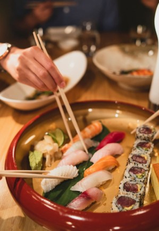 Person using chopsticks to pick up sushi from a wooden boat platter containing assorted nigiri and specialty rolls