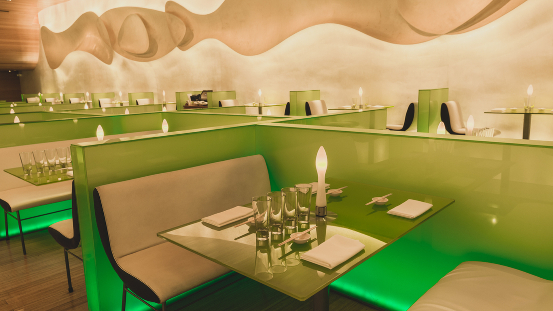 Modern restaurant with green-lit booths, light-colored curved benches, and abstract mural on the wall.