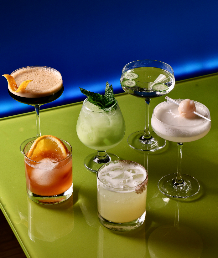 Six colorful cocktails in various glassware, including a coupe, rocks glass, and margarita glass, displayed on a green surface against a blue background.