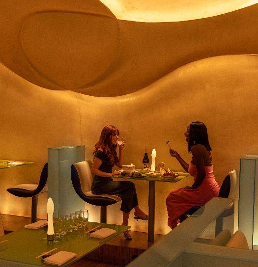 Two guests dining at a round table in a softly glowing, sculptural amber dining room.