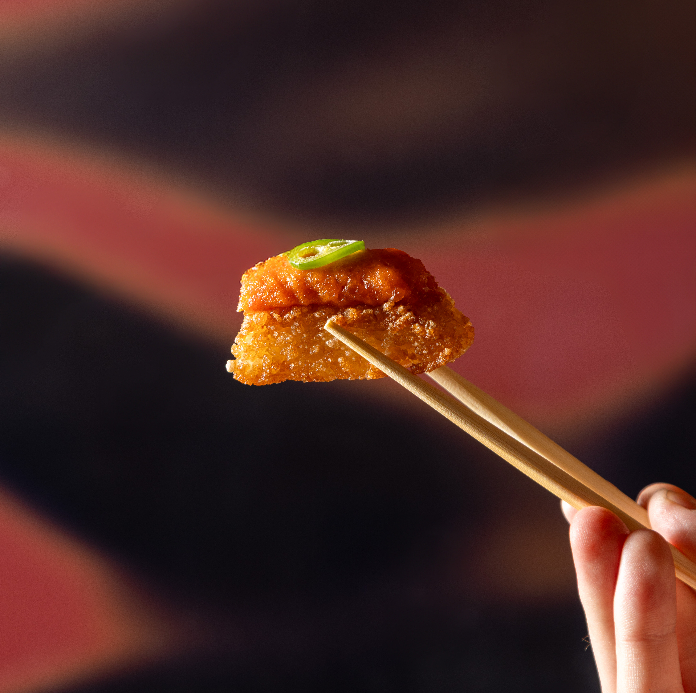 A close-up of chopsticks holding a crispy bite topped with bright orange sauce and a slice of green chili.