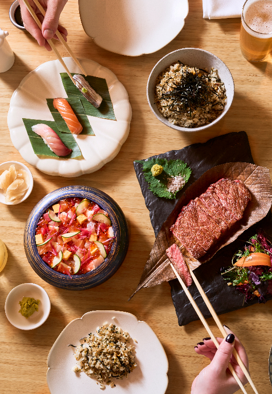 An overhead view of a Japanese-inspired meal with sushi, rice, seafood, steak, and small plates arranged on a wooden table.