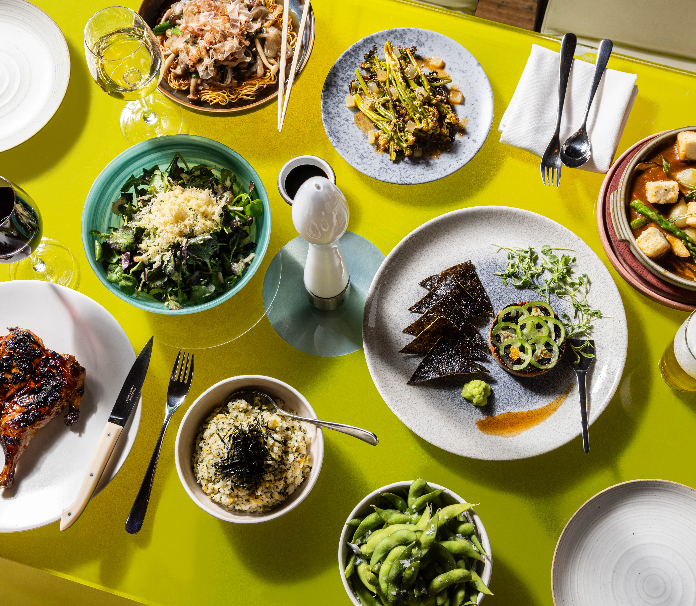 A colorful tabletop spread of Japanese-inspired dishes, including greens, rice, seaweed, edamame, noodles, and grilled items.